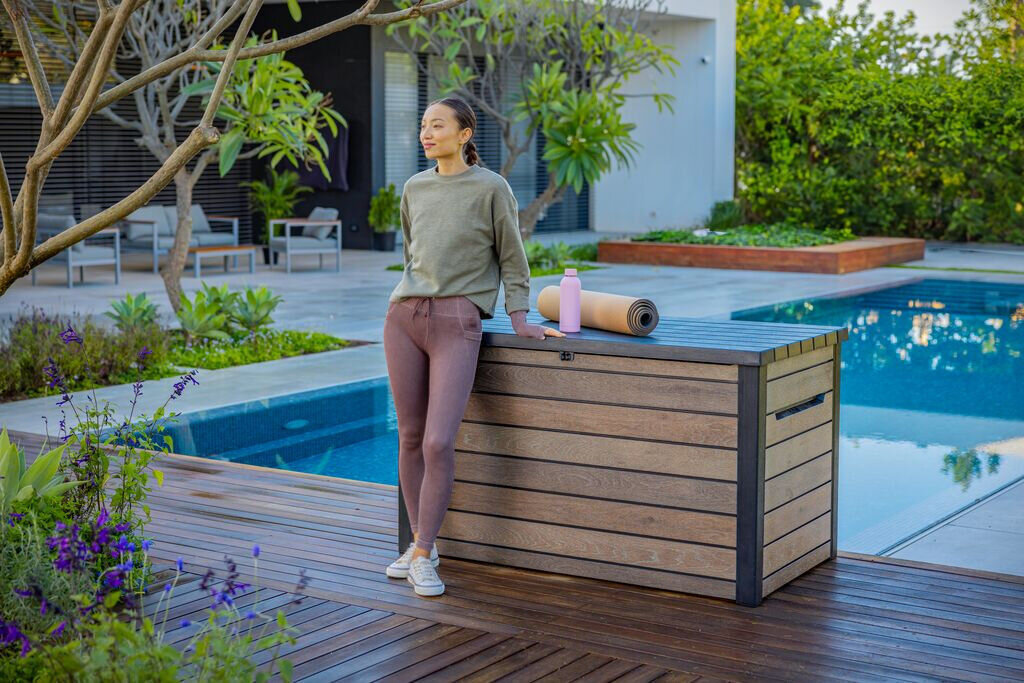 Na drewnianym tarasie przy basenie stoi brązowa skrzynia ogrodowa z ciemnymi krawędziami. Na skrzyni leży mata do ćwiczeń i różowa butelka.