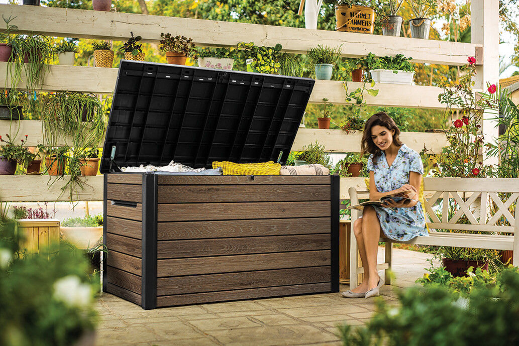 Na zdjęciu znajduje się brązowa skrzynia ogrodowa z otwartą pokrywą, wypełniona różnymi przedmiotami. Obok skrzyni siedzi osoba na ławce, otoczona roślinami w doniczkach.