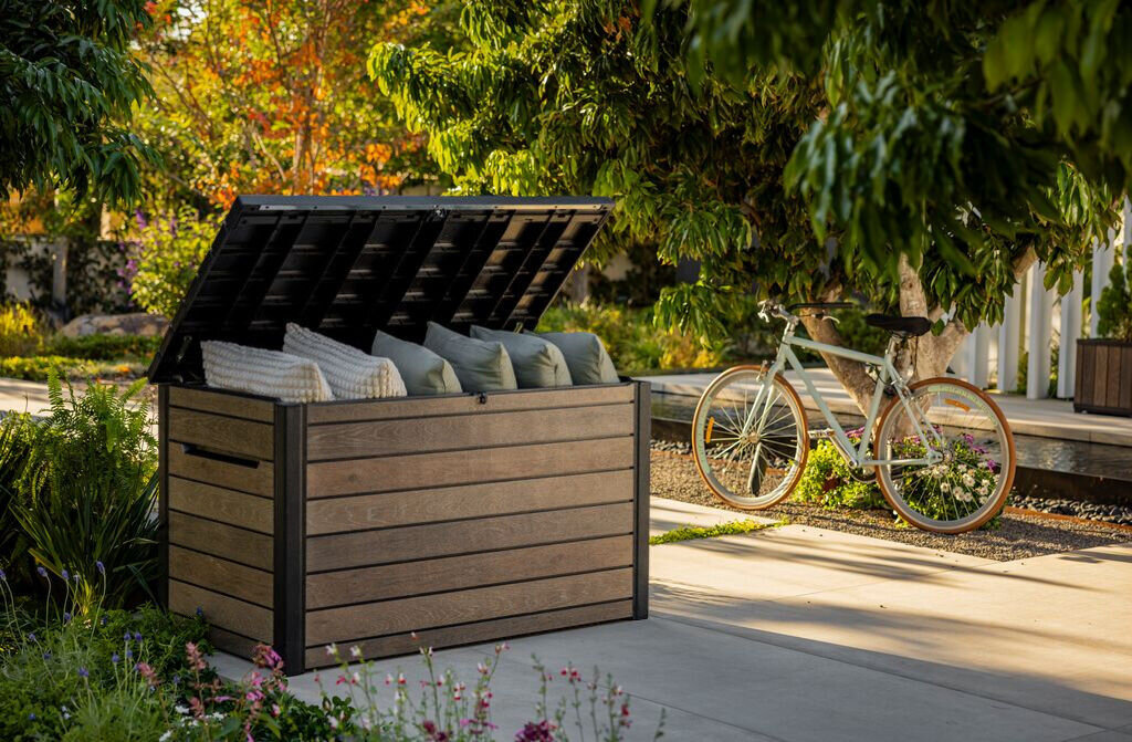 Na zdjęciu znajduje się brązowa skrzynia ogrodowa z otwartą pokrywą, wypełniona poduszkami. W tle widać rower oraz roślinność ogrodową.