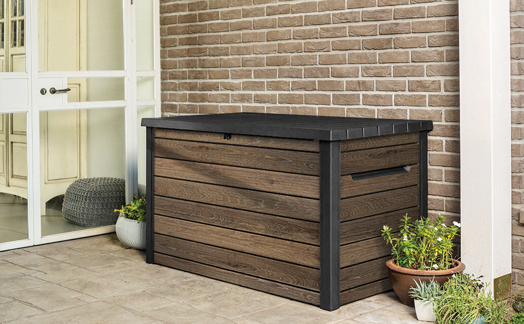 Na obrazie widoczna jest brązowa skrzynia ogrodowa z ciemnym wiekiem, ustawiona na tarasie przy ścianie z cegieł. Obok skrzyni znajdują się doniczki z roślinami.