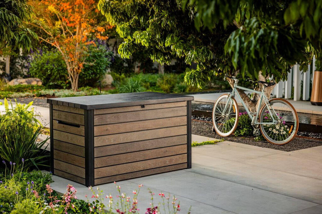 Na zdjęciu znajduje się brązowa skrzynia ogrodowa z poziomymi panelami, ustawiona na tarasie w otoczeniu zieleni. W tle widoczny jest rower oparty o drzewo.