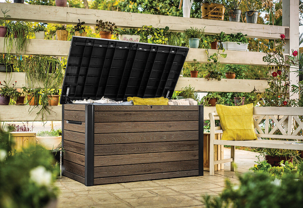 Brązowa skrzynia ogrodowa z otwartą pokrywą stoi na tarasie, otoczona roślinami w doniczkach. Obok znajduje się biała ławka z żółtą poduszką.