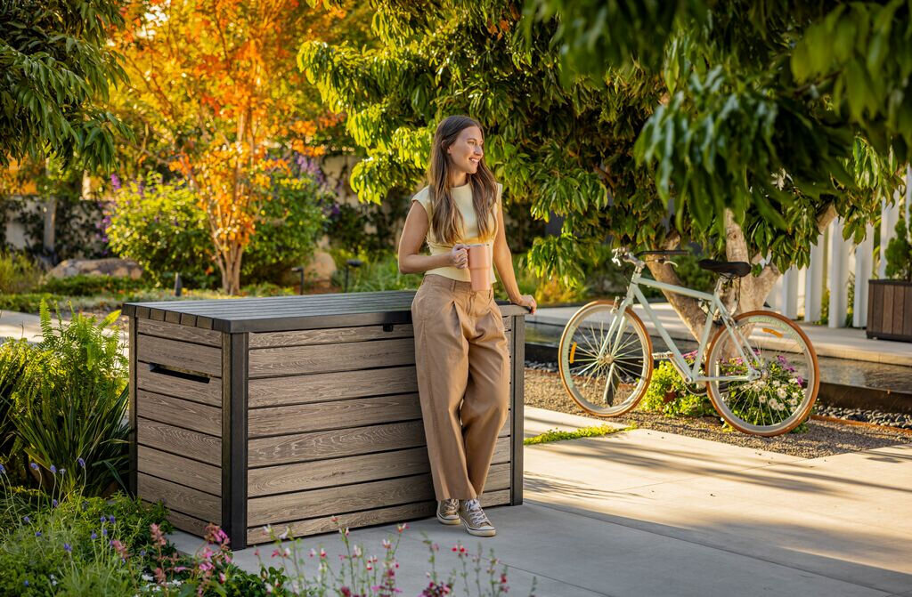 Na zdjęciu znajduje się szaro-brązowa skrzynia ogrodowa z drewnianym wykończeniem, obok której stoi osoba trzymająca kubek. W tle widoczny jest rower oraz bujna roślinność ogrodowa.