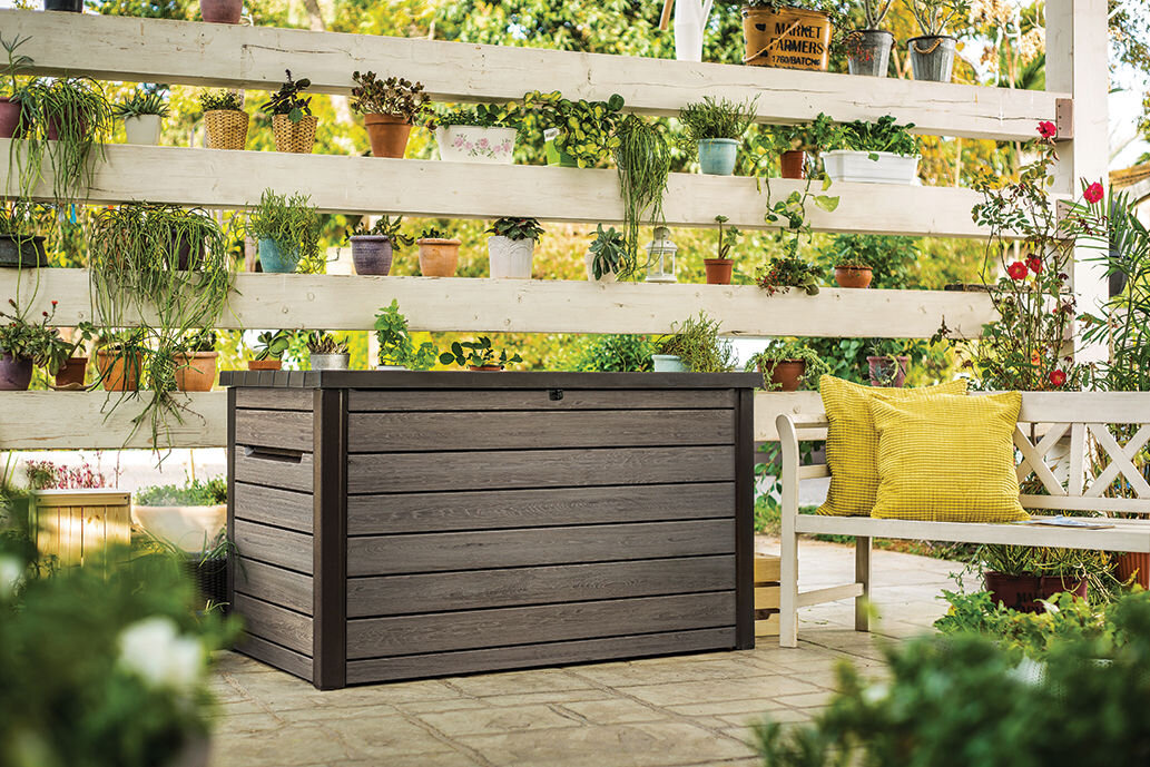 Na tarasie znajduje się szaro-brązowa skrzynia ogrodowa o drewnopodobnej fakturze. W tle widać drewnianą konstrukcję z doniczkami pełnymi roślin.