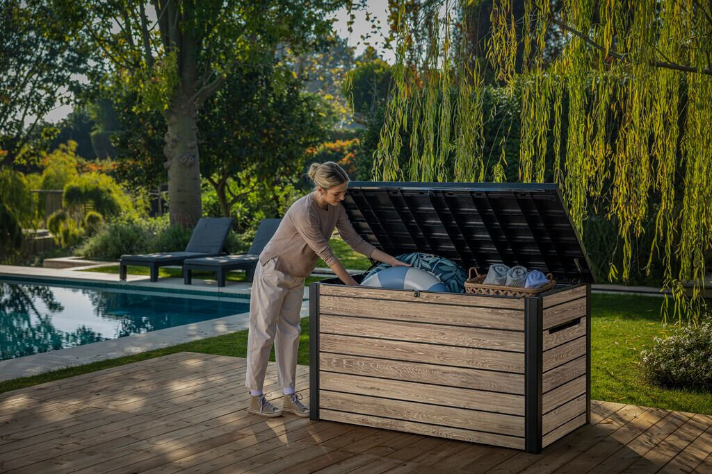 Osoba stoi przy otwartej skrzyni ogrodowej, wkładając do niej poduszki i koce. Skrzynia ma szaro-brązowy kolor i znajduje się na drewnianym tarasie obok basenu, otoczona zielenią.