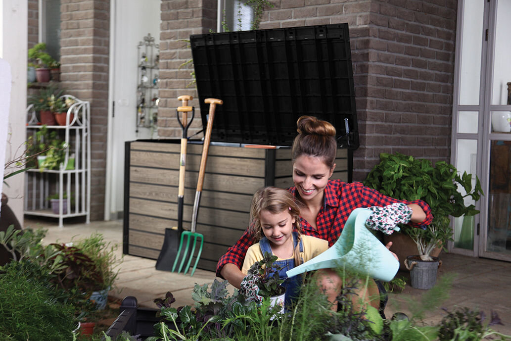 Na obrazie widać otwartą skrzynię ogrodową w kolorze szaro-brązowym, obok której znajdują się narzędzia ogrodnicze. W tle widoczne są rośliny doniczkowe i ceglana ściana.