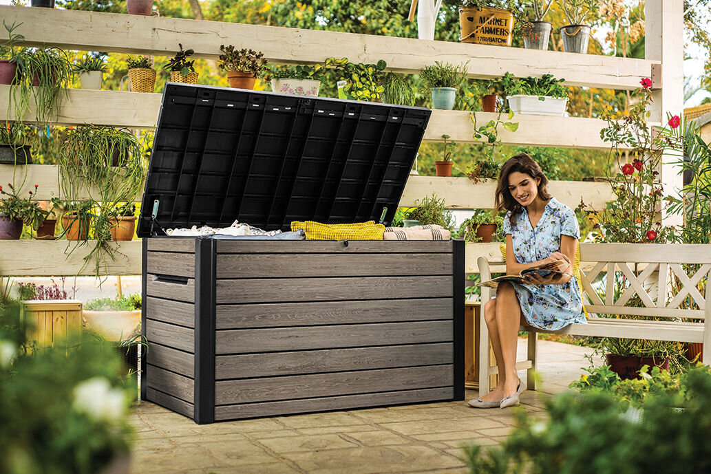 Na zdjęciu znajduje się otwarta skrzynia ogrodowa w kolorze szaro-brązowym, ustawiona na tarasie otoczonym roślinami w doniczkach. Obok skrzyni siedzi osoba na ławce, trzymająca książkę.