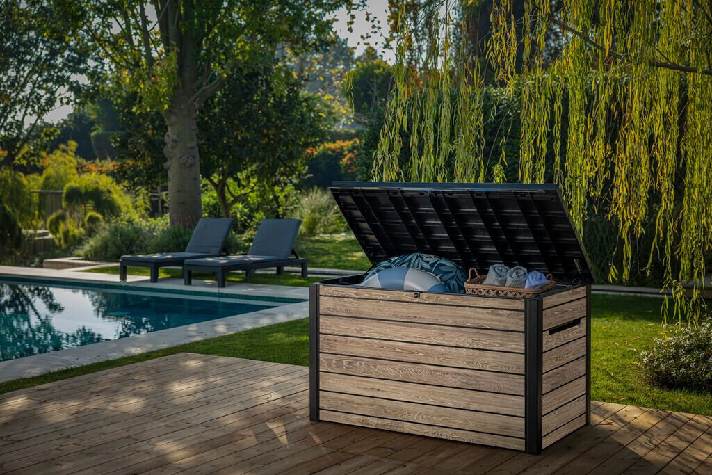 Otwarta skrzynia ogrodowa w kolorze szaro-brązowym stoi na drewnianym tarasie, wypełniona różnymi przedmiotami. W tle widoczny jest basen, leżaki oraz zieleń ogrodu.