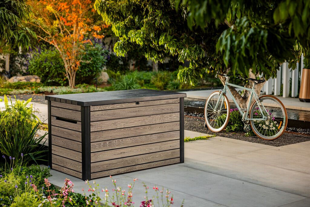 Na obrazie znajduje się szaro-brązowa skrzynia ogrodowa z drewnopodobnym wykończeniem, ustawiona na tarasie w otoczeniu zieleni. W tle widoczny jest rower oparty o drzewo.