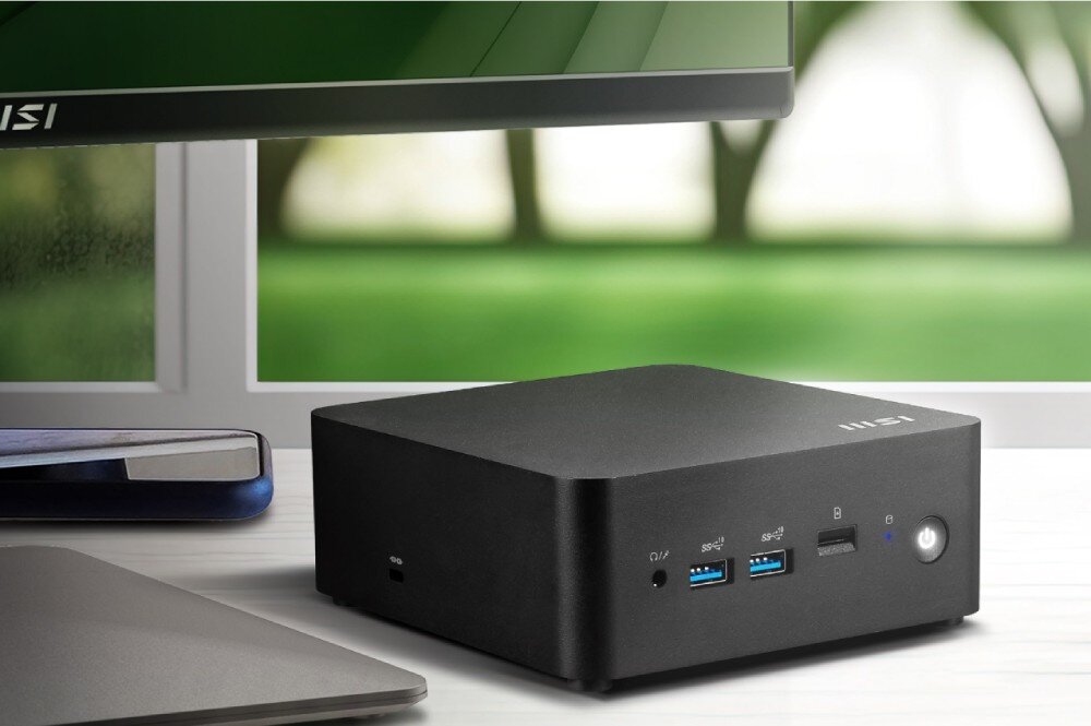 Komputer MSI Cubi NUC 13MQ-062EU Niewielki komputer w czarnej obudowie ustawiony na biurku obok monitora, laptopa i smartfona przy oknie z widokiem na zieleń, opis
