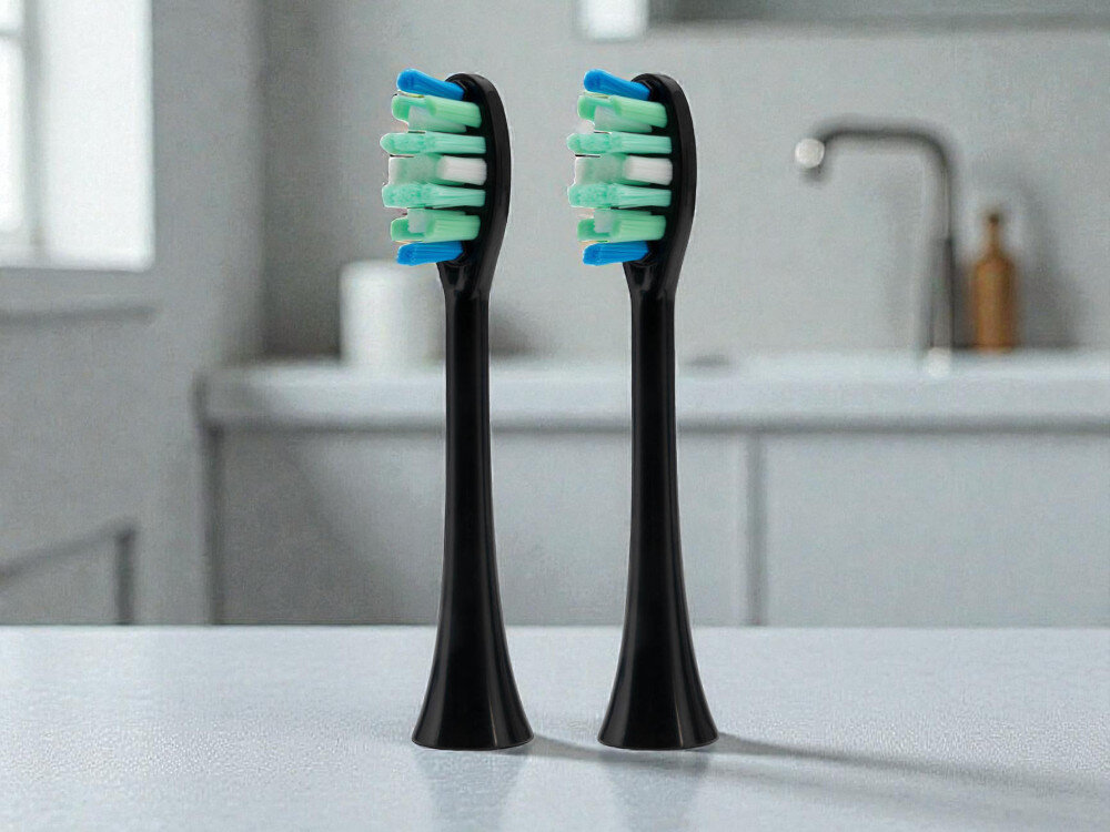 Oryginalna końcówka szczoteczki GÖTZE & JENSEN ASTH956 Daily Care średnie włosie do usuwania płytki nazębnej kompatybilna ze szczoteczką soniczną ST955 Dwie czarne końcówki do szczoteczki sonicznej z kolorowym włosiem stojące na jasnym blacie w nowoczesnej łazience.