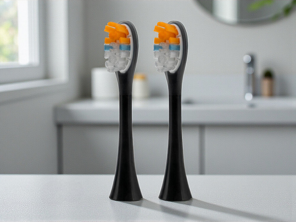 Oryginalna końcówka szczoteczki GÖTZE & JENSEN ASTH957 Daily Care średnie włosie do usuwania płytki nazębnej kompatybilna ze szczoteczką soniczną ST955 Dwie czarne końcówki do szczoteczki sonicznej z kolorowym włosiem stojące na jasnym blacie w nowoczesnej łazience.