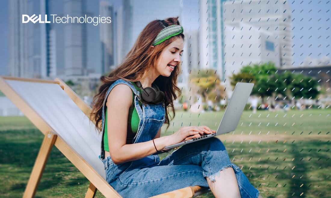 Kobieta pracująca na laptopie Dell Inspiron Plus 7440 na leżaku w parku, słuchawki na szyi, w tle wieżowce i logo Dell Technologies.