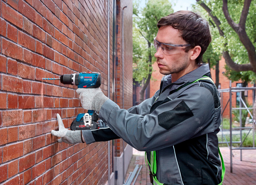 Wiertarko-wkrętarka BOSCH Professional GSB 18V-65 06019N3301 budynek z cegły osoba wierci otwór w ścianie w tle drzewa rusztowanie chodnik funkcja udaru z maksymalną liczbą do 27000 udarów/min szybkie, pulsacyjne ruchy wspierają pracę wiertła otwory powstają sprawniej skraca się czas pracy i zmniejsza nacisk potrzebny do prowadzenia narzędzia