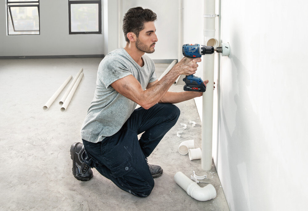 Wiertarko-wkrętarka BOSCH Professional GSR 18V-65 06019N3201 pomieszczenie biała ściana osoba trzyma wiertarko-wkrętarkę w dłoniach wierci dziurę w ścianie w tle dwa małe okna waga około 1,06 kg bez akumulatora smukła dobrze wyprofilowana rękojeść środkowa pokrycie Softgrip z antypoślizgowego miękkiego materiału długość korpusu wynosząca 166 mm stabilność chwytu ogranicza ślizganie się dłoni