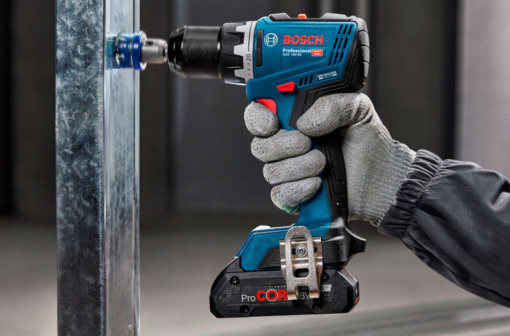 Wiertarko-wkrętarka BOSCH Professional GSR 18V-65 06019N3201 pomieszczenie metalowe profile osoba trzyma wiertarko-wkrętarkę w dłoniach wierci dziurę w profilu metalowy zaczep można zawiesić na pasku lub narzędziowym pasie roboczym ułatwia przenoszenie i szybki dostęp zwiększa komfort i tempo pracy