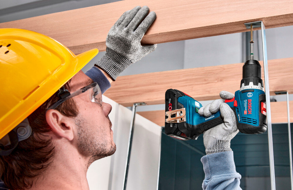 Wiertarko-wkrętarka BOSCH Professional GSR 18V-65 06019N3200 pomieszczenie drewniana konstrukcja osoba trzyma wiertarko-wkrętarkę w dłoniach przykręca metalowy hak do konstrukcji widok od tyłu wkręcanie wkrętów w drewnie wiercenie w metalu jedno urządzenie pozwala wykonać więcej prac szybciej i przy mniejszym bałaganie