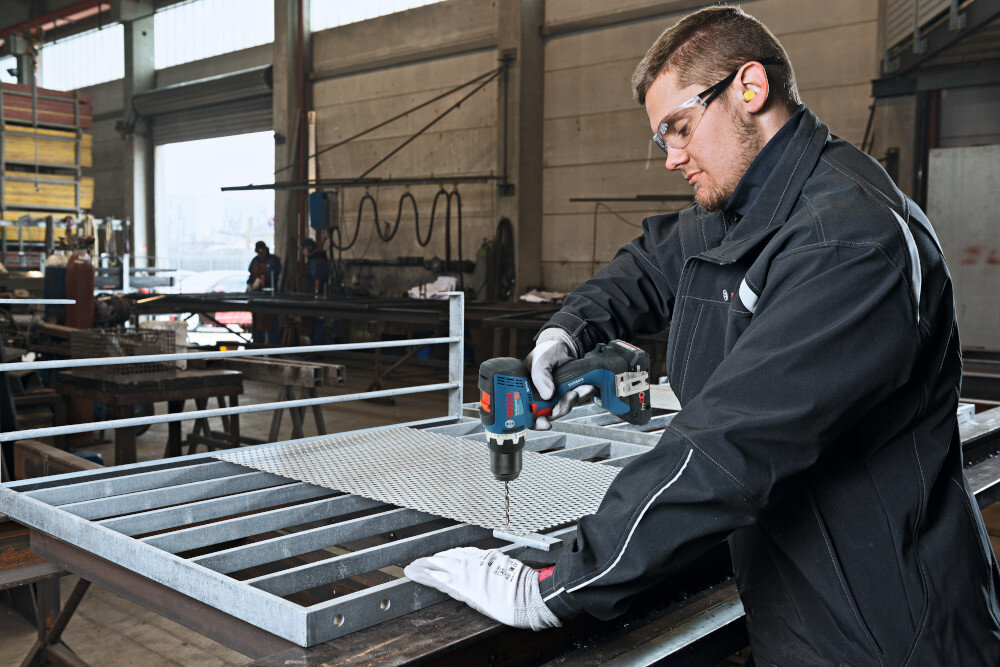 Wiertarko-wkrętarka BOSCH Professional GSR 18V-65 06019N3200 pomieszczenie warsztat metalowa konstrukcja osoba trzyma wiertarko-wkrętarkę w dłoniach wierci otwór w konstrukcji w tle duże drzwi szybkozaciskowy uchwyt wiertarski 13 mm sprawna wymiana wierteł i bitów bez użycia dodatkowych kluczy zakres mocowania 1,5–13 mm cienkie wiertła do precyzyjnych otworów większe akcesoria do cięższych prac