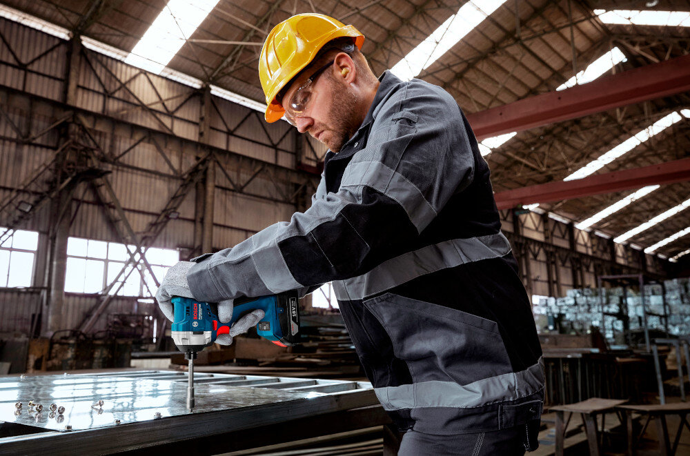 Klucz udarowy BOSCH Professiona GDR 18V-215 06019N2000 warsztat osoba wkręca śruby w metalową blachę obudowa odporna na uszkodzenia ergonomiczny kształt rękojeści antypoślizgowa powłoka waga około 1,0 kg bez akumulatorem większy komfort i lepsza kontrola narzędzia