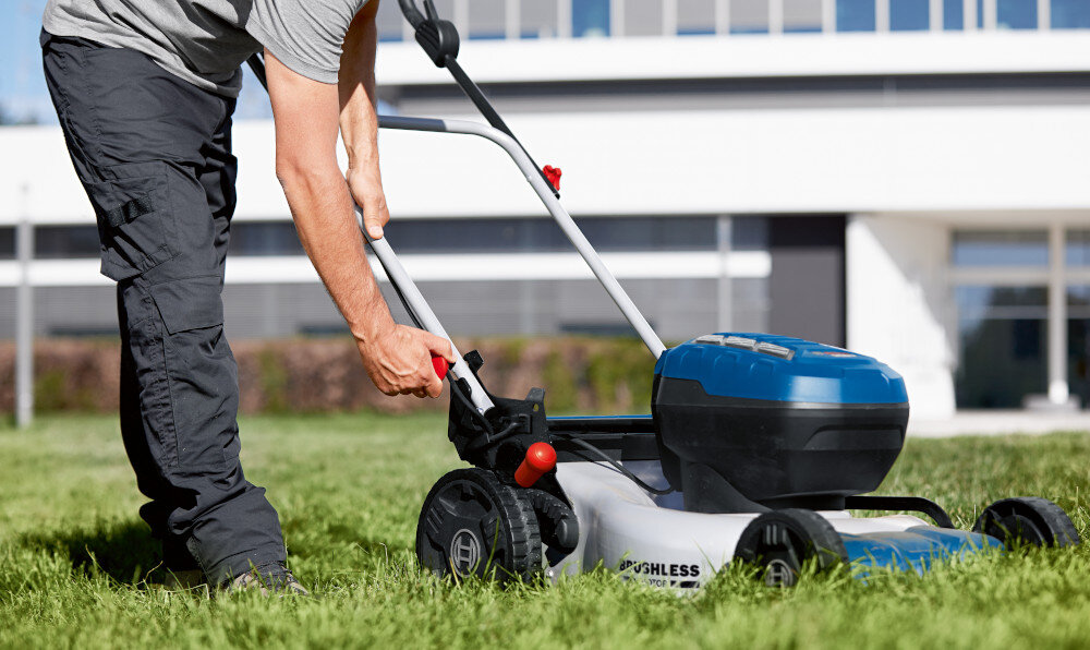Kosiarka akumulatorowa BOSCH Professional GRA 18V2-46 06008C8000 trawnik w tle budynek osoba pochyla się przy kosiarce stalowy nóż obrotowy o szerokości 46 cm szybkie i efektywne koszenie redukuje liczbę przejść równomierne i czyste cięcie