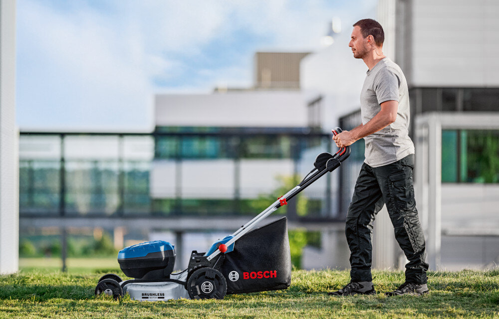 Kosiarka akumulatorowa BOSCH Professional GRA 18V2-46 06008C8000 ogród w tle budynek osoba kosi trawnik uchwyt z miękkim wykończeniem komfort pracy zapobiega nadmiernemu zmęczeniu dłoni dwustopniowa regulacja wysokości kabłąk pełni funkcję dodatkowego uchwytu pewne prowadzenie kosiarki kontrolę nad kierunkiem jazdy ułatwia manewrowanie