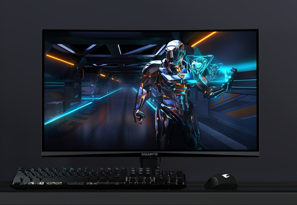 Monitor GIGABYTE GS27QCA Monitor wyświetlający futurystyczną postać robota w błyszczącej zbroi, stojącą w oświetlonym korytarzu o niebiesko-pomarańczowej kolorystyce. Pod monitorem znajduje się klawiatura i myszka, opis