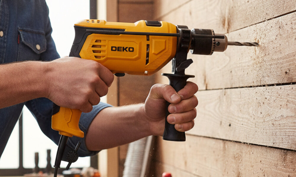 Wiertarka udarowa DEKO DKID13Y65 drewniana ściana osoba wierci otwór w ścianie zbliżenie na uchwyt skosem ergonomiczny uchwyt materiał antypoślizgowy stabilność prowadzenia urządzenia dodatkowa rękojeść precyzja i bezpieczeństwo waga około 1,615 kg
