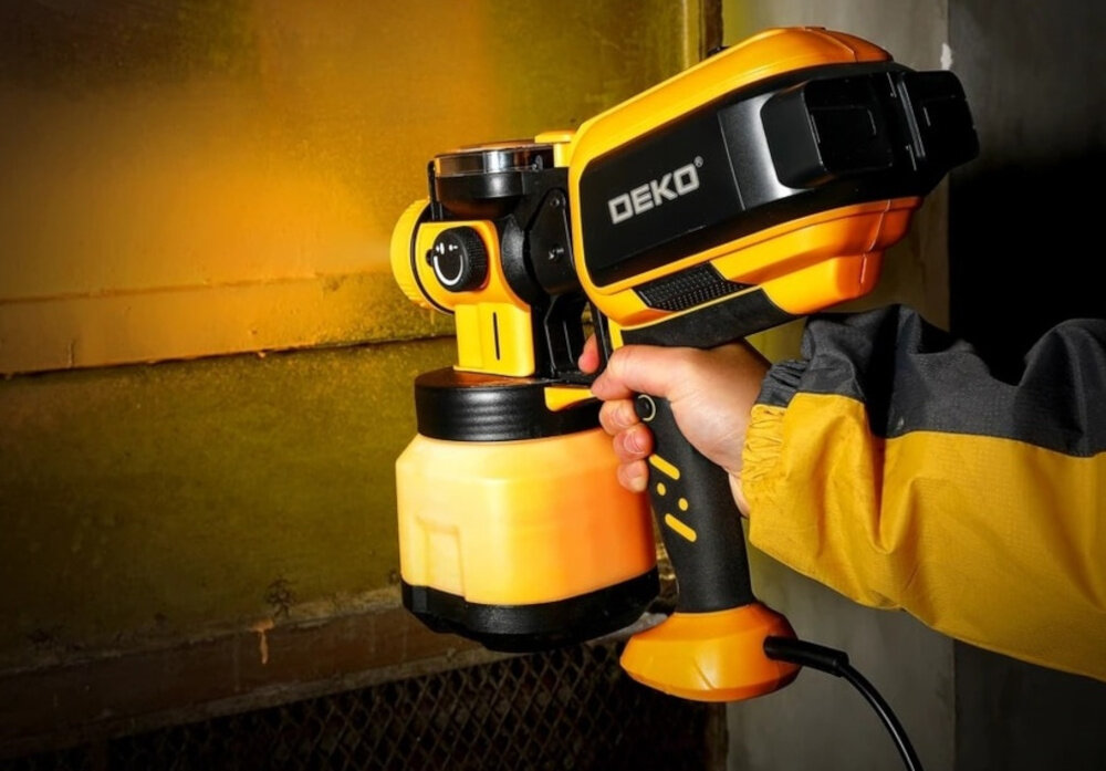 Pistolet natryskowy DEKO DKSG60K2 1200 ml osoba trzyma pistolet w dłoni maluje ścianę żółta farba technologia HVLP oszczędność farby nawet do 30% praca na niskim ciśnieniu ogranicza ilość rozprysków czystość miejsca pracy