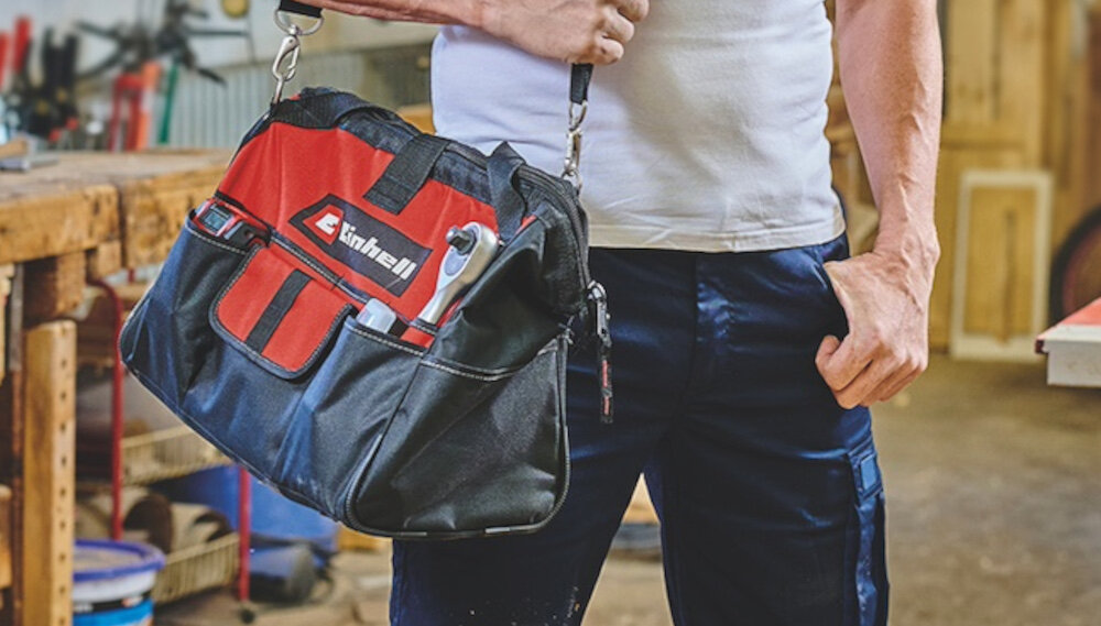 Torba narzędziowa EINHELL 4530074 pomieszczenie warsztat osoba niesie torbę na ramieniu w tle stół warsztatowy drewniane elementy z wytrzymałego nylonu lepiej opiera się przetarciom i rozciąganiu wzmocnione dno szwy i łączenia materiału zostały dodatkowo wzmocnione torba utrzymuje kształt