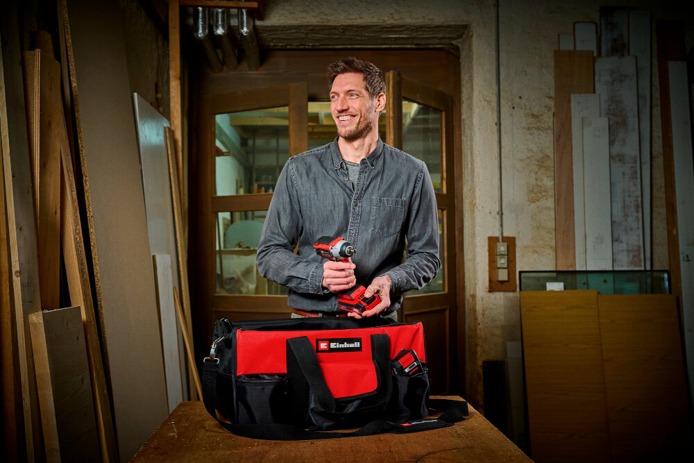 Torba narzędziowa EINHELL 4530078 pomieszczenie warsztat torba ustawiona na drewnianym stole osoba wyciąga z niej narzędzia w tle drzwi drewniane elementy wielokomorowa konstrukcja udźwig do 25 kg liczne kieszenie i przegródki specjalne wewnętrzne przegrody miejsca na akumulatory PXC 