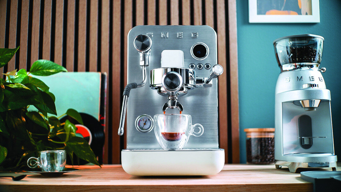 Na drewnianym blacie stoi srebrny ekspres do kawy z logo \'SMEG\', obok którego znajduje się szklanka z kawą. W tle widoczny jest młynek do kawy oraz roślina doniczkowa.