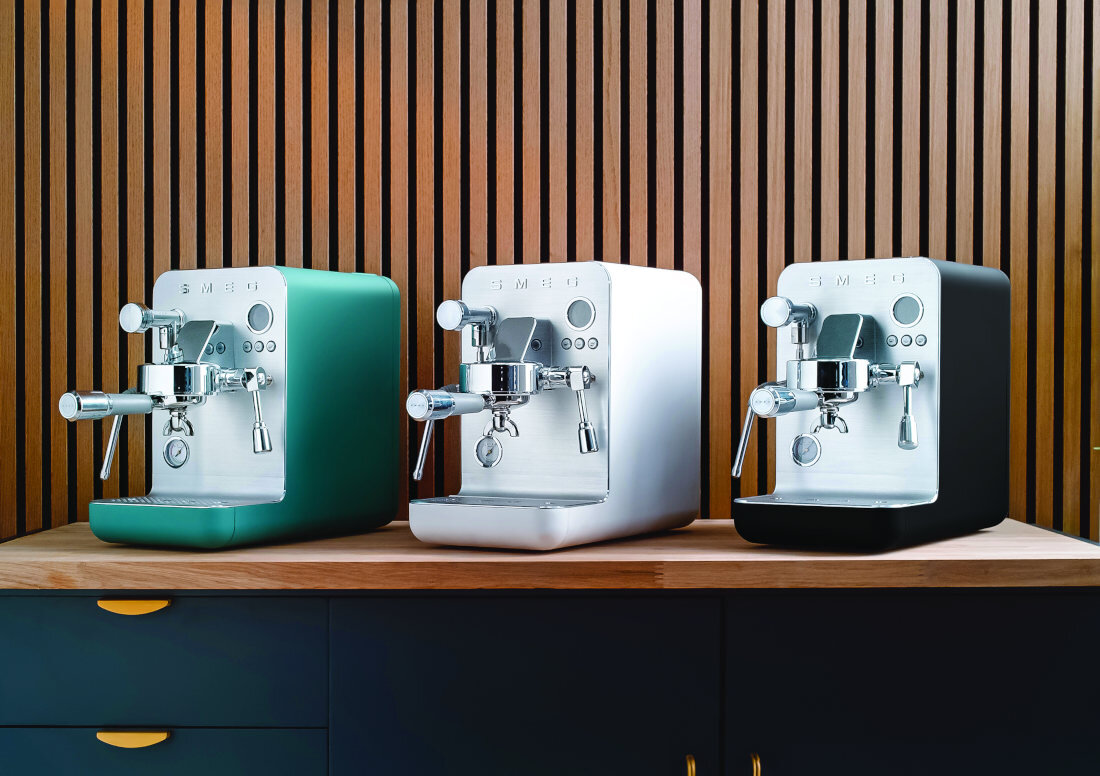 Trzy ekspresy do kawy SMEG w kolorach zielonym, białym i czarnym stoją na drewnianym blacie przed ścianą z pionowych drewnianych listew. Każdy ekspres ma metaliczne wykończenie z przodu i widoczne pokrętła oraz dysze.