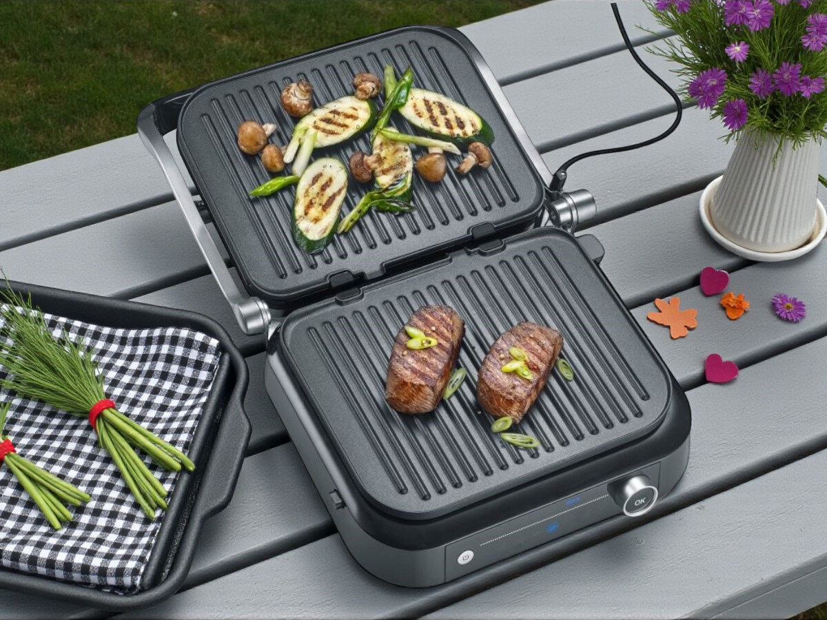 Grill elektryczny SEVERIN KG 2395, powierzchnia grillowania, grill kontaktowy, grill stołowy, moc 2000 W, Grill elektryczny ustawiony na blacie kuchennym, uchylona pokrywa, na dolnej płycie grillowane mięso i zielone papryczki shishito. W tle dwójka kucharzy ubranych w białe koszule i czarne fartuchy, pracujących na profesjonalnej kuchni z ceglaną ścianą oraz półkami z naczyniami.