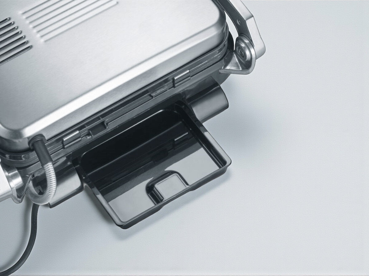 Grill elektryczny SEVERIN KG 2395, 7 programów, czujnik grubości, timer, sygnał dźwiękowy, Zbliżenie na górną część grilla elektrycznego w kolorze stalowym, ustawioną na jasnym blacie. Widoczna wyjmowana czarna tacka ociekowa na tłuszcz i przewód z metalowym oplotem, podkreślające łatwość czyszczenia i utrzymania urządzenia w czystości.