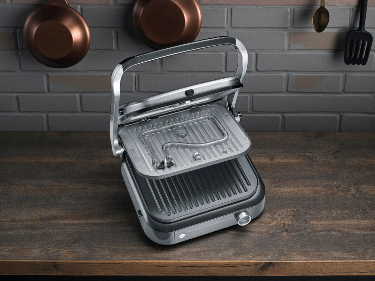 Grill elektryczny SEVERIN KG 2395, powłoka ceramiczna, płyty wyjmowane, łatwe czyszczenie, tacka na tłuszcz, Otwarty grill elektryczny prezentowany na drewnianym blacie kuchennym, widok z góry. Pokrywa uniesiona, płyty grillowe czyste, widoczne elementy grzewcze od spodu górnej płyty. W tle ściana z cegły i wiszące miedziane garnki oraz kuchenne akcesoria.