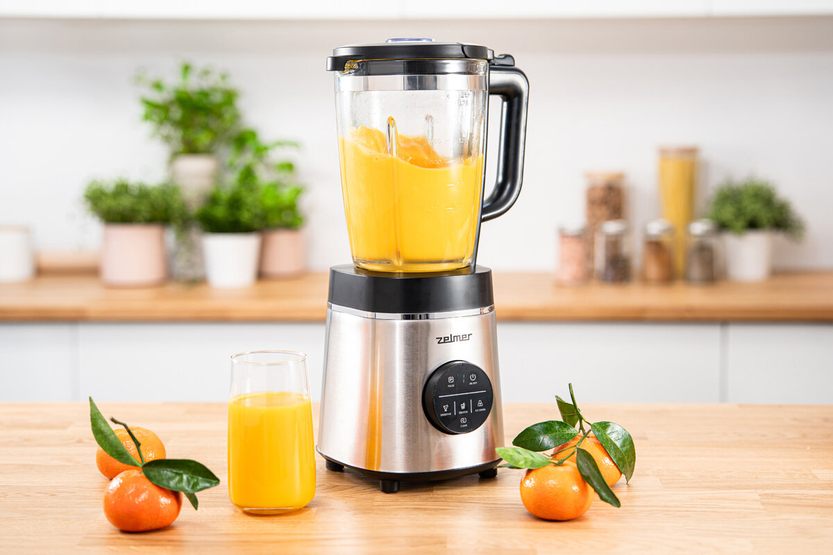 Na blacie kuchennym stoi blender kielichowy z logo \'Zelmer\', wypełniony pomarańczowym płynem. Obok znajduje się szklanka z sokiem i kilka mandarynek z liśćmi.