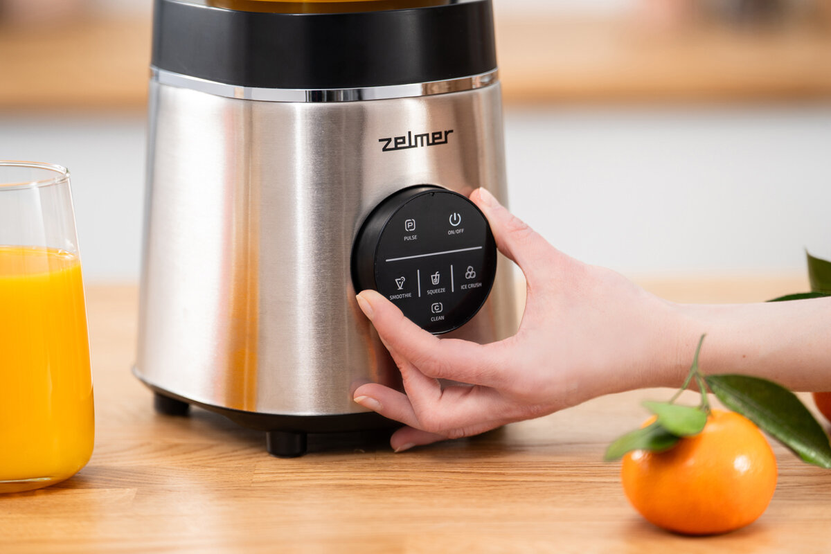 Osoba obsługuje blender kielichowy marki Zelmer, który ma srebrno-czarną obudowę i okrągły panel sterowania z przyciskami. Obok znajduje się szklanka z sokiem pomarańczowym i mandarynka.