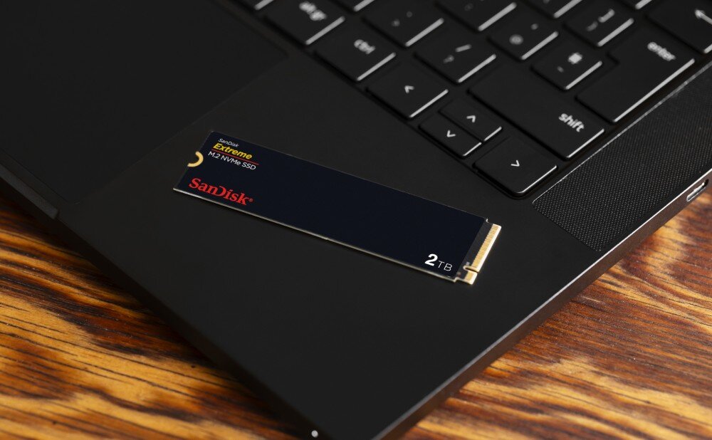 Dysk SANDISK Extreme Mały dysk SSD w formacie M.2 leży na obudowie czarnego laptopa umieszczonego na drewnianym blacie, opis