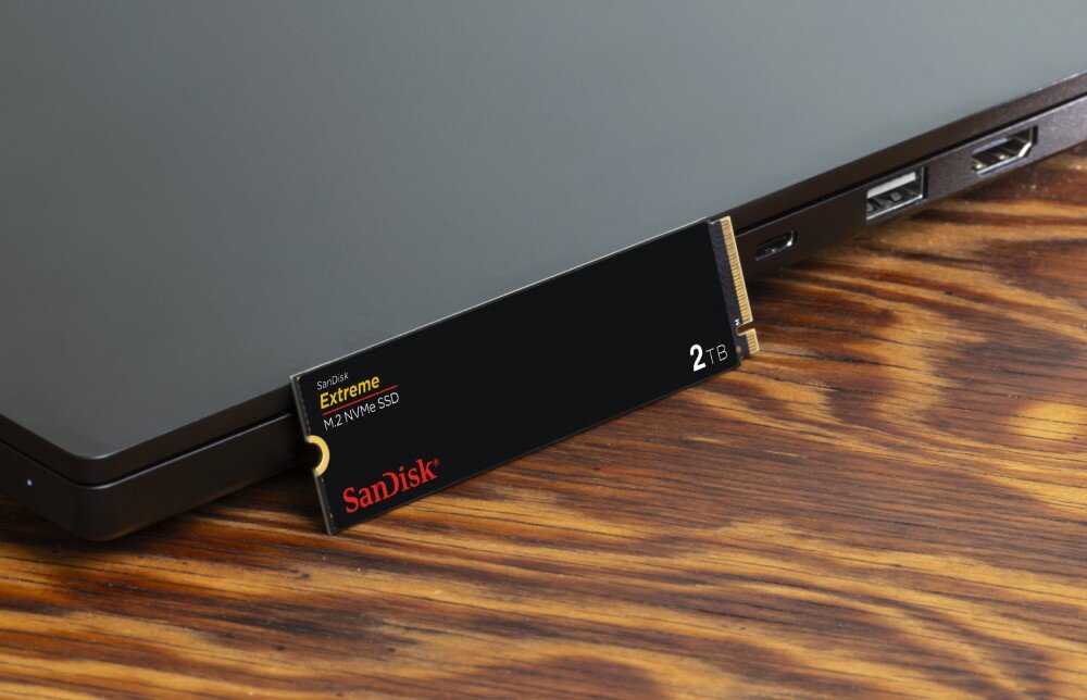 Dysk SANDISK Extreme Dysk SSD M.2 częściowo wsunięty w złącze laptopa stojącego na drewnianym blacie, wydajność, odczyt, zapis