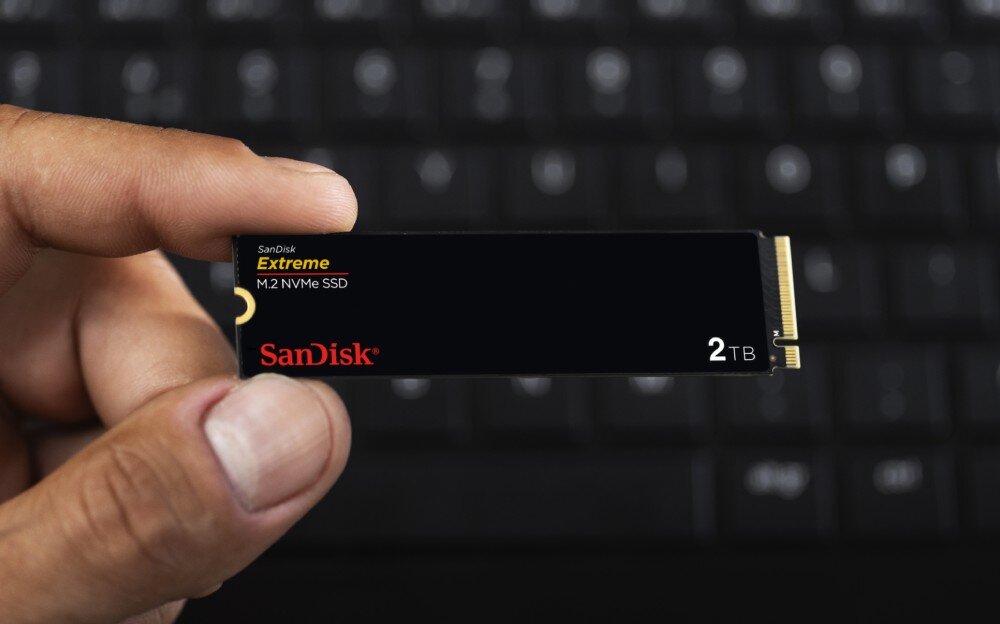 Dysk SANDISK Extreme Dłoń trzyma mały dysk SSD M.2, w tle rozmazana klawiatura laptopa, gniazdo, montaż, oprogramowanie