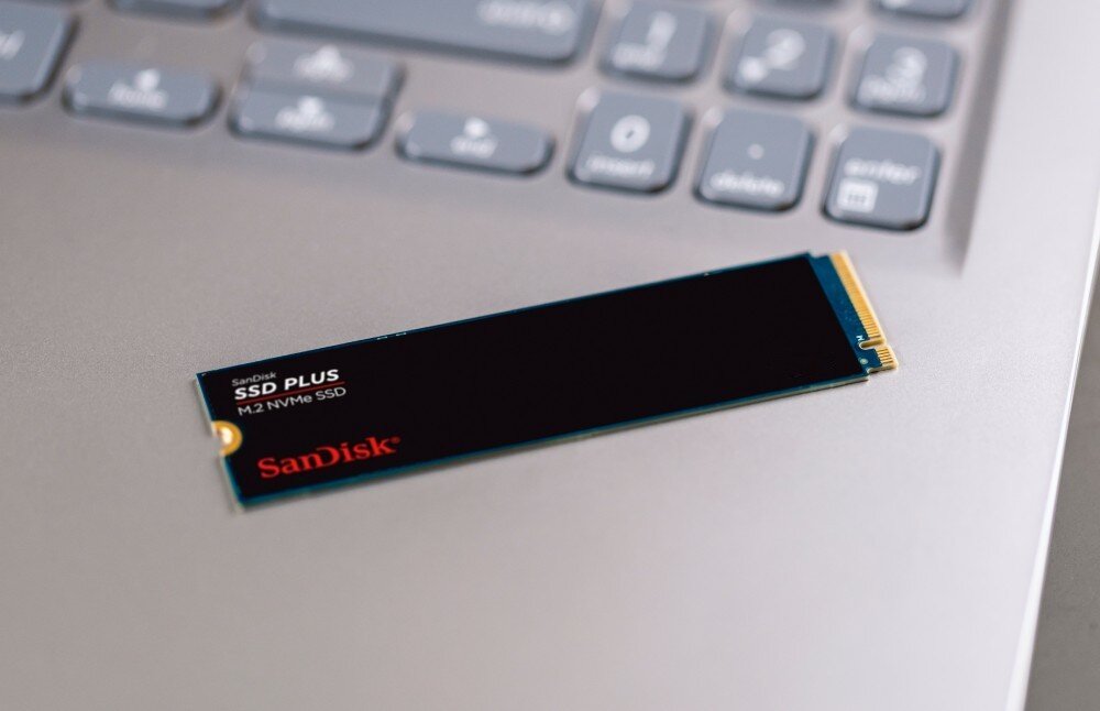 Dysk SANDISK Plus Wąski dysk w formacie M.2 leży na srebrnym laptopie obok klawiatury, opis