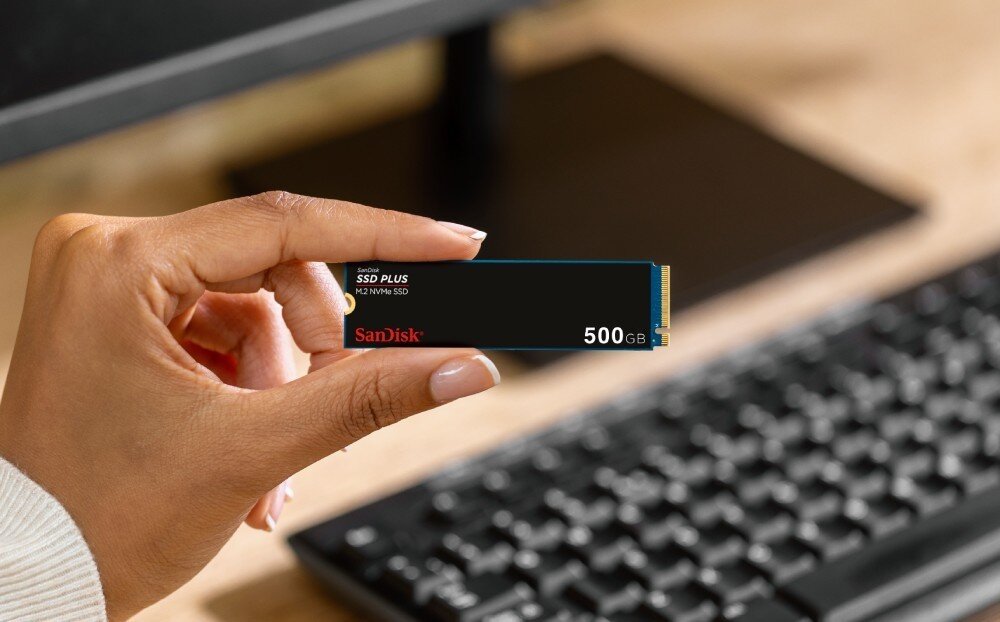 Dysk SANDISK Plus Osoba trzyma w palcach wąski dysk M.2, w tle widoczna klawiatura i monitor na biurku, przeznaczenie, cechy