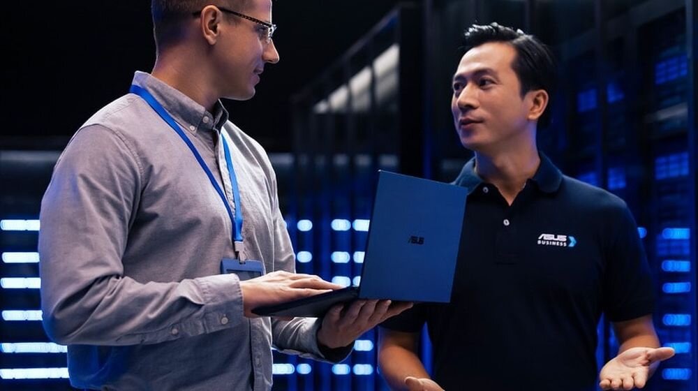 Dwóch specjalistów IT na konferencji technologicznej z laptopem ASUS ExpertBook B5, mężczyzna w szarej kamizelce z identyfikatorem konferencyjnym i azjatycki przedstawiciel ASUS w czarnej koszulce polo, certyfikat MIL-STD-810H zapewniający wytrzymałość na trudne warunki dla profesjonalistów biznesowych - MIL-STD-810H