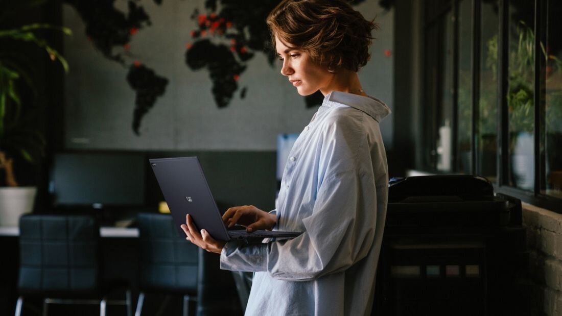 Kobieta w białej koszuli pracująca na laptopie ASUS ExpertBook B5 w stylowym wnętrzu z mapą świata na ścianie, bateria 63 Wh do długiej pracy mobilnej, eleganckie biuro z roślinami i ciemnymi meblami, rozwiązanie dla osób pracujących w podróży i profesjonalistów  - Bateria o pojemności 63 Wh  