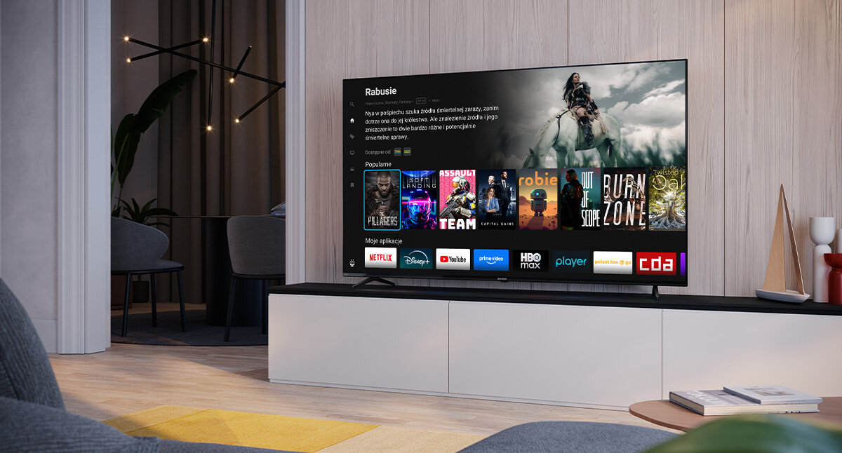 Telewizor wyświetla ekran główny z aplikacjami takimi jak Netflix, Disney+, YouTube, Prime Video, HBO Max, Player, Polsat Box Go i CDA. W tle widoczny jest nowoczesny salon z rośliną i dekoracjami.