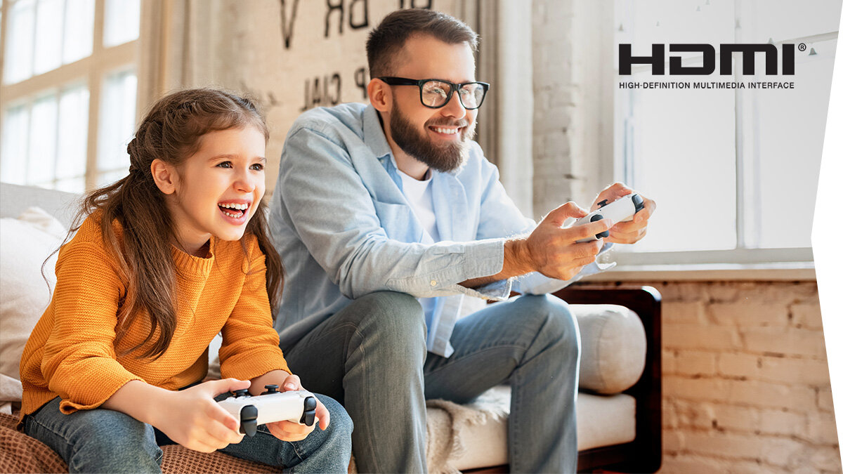 Dwie osoby siedzą na kanapie, trzymając kontrolery do gier. W prawym górnym rogu widoczne jest logo HDMI z napisem \'High-Definition Multimedia Interface\'.