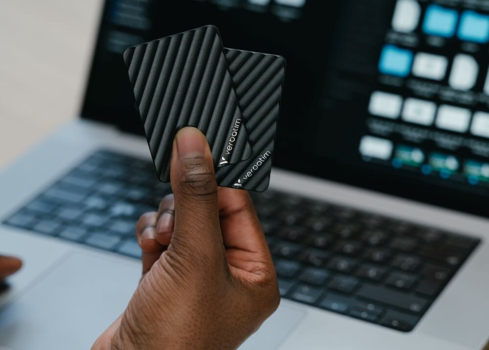 Dysk VERBATIM Store n Go Mini Stripe 2TB SSD Dłoń trzymająca dwa czarne urządzenia wielkości karty z ukośnymi żłobieniami na tle laptopa, opis