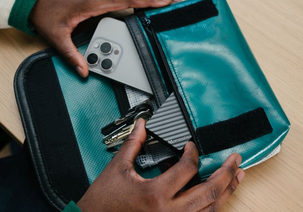 Dysk VERBATIM Store n Go Mini Stripe 2TB SSD Ręce wkładają czarne urządzenie z żłobieniami do zielonej torby obok smartfona i pęku kluczy, oprogramowanie, kompatybilność