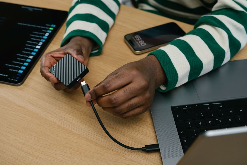Dysk VERBATIM Store n Go Mini Stripe 1TB SSD Osoba w zielono-białej bluzie podłącza czarne urządzenie z żłobieniami do laptopa kablem USB-C, wydajność, interfejs
