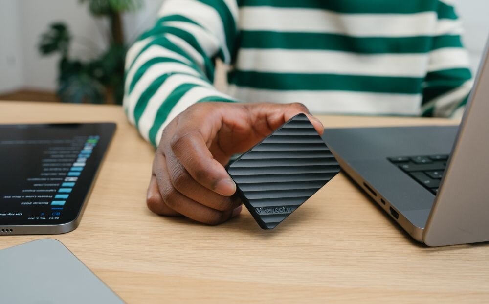Dysk VERBATIM Store n Go Mini Stripe 512GB SSD Osoba w zielono-białej bluzie trzyma czarne urządzenie z żłobieniami obok laptopa na biurku, wymiary, waga, konstrukcja